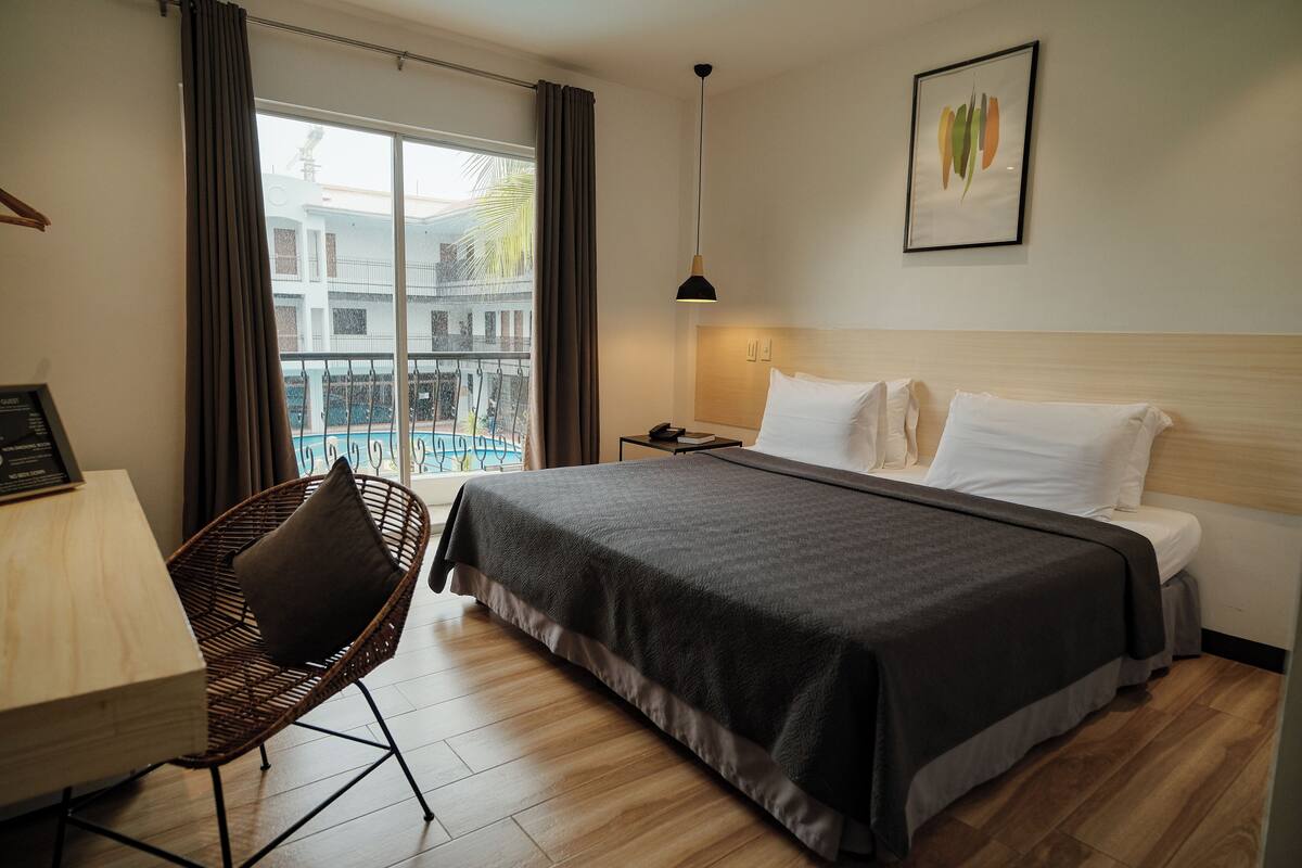 A hotel room with a bed, a chair, a nightstand, a lamp, a window with curtains, and a view of buildings outside.