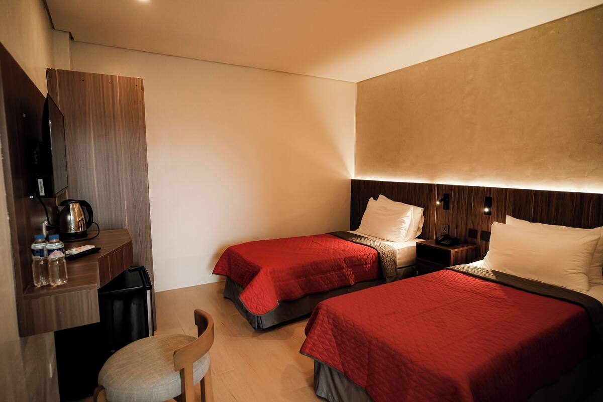 A hotel room with two beds, a desk with a kettle, a chair, and a TV.