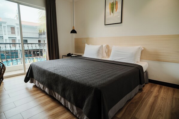Ein Hotelzimmer mit einem großen Bett, einem hölzernen Kopfteil, einer schwarzen Pendelleuchte und Blick auf einen Pool und Gebäude durch ein großes Fenster.
