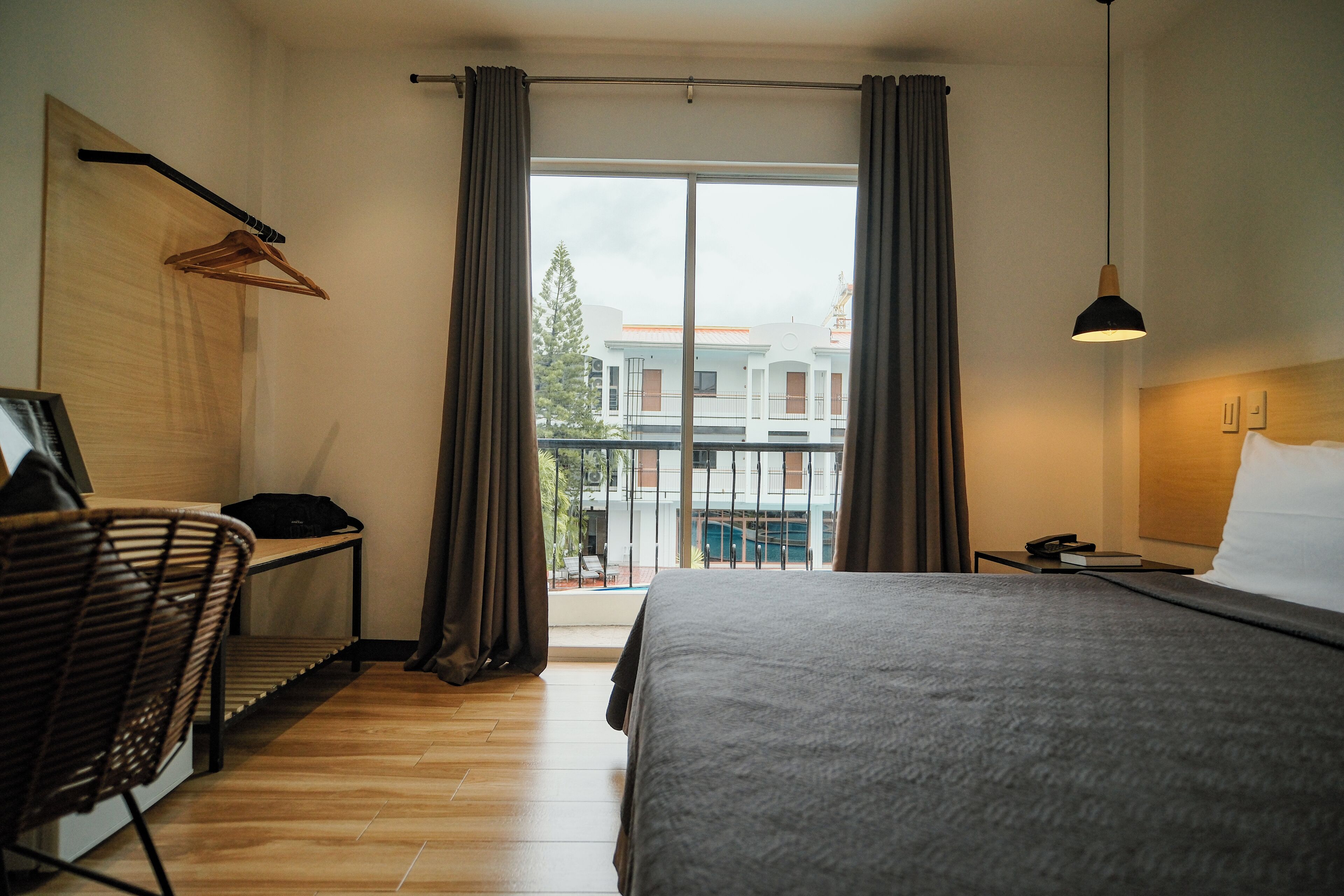 A hotel room with a bed, a chair, a desk, a hanging coat rack, and a view of a building and trees through a large window.