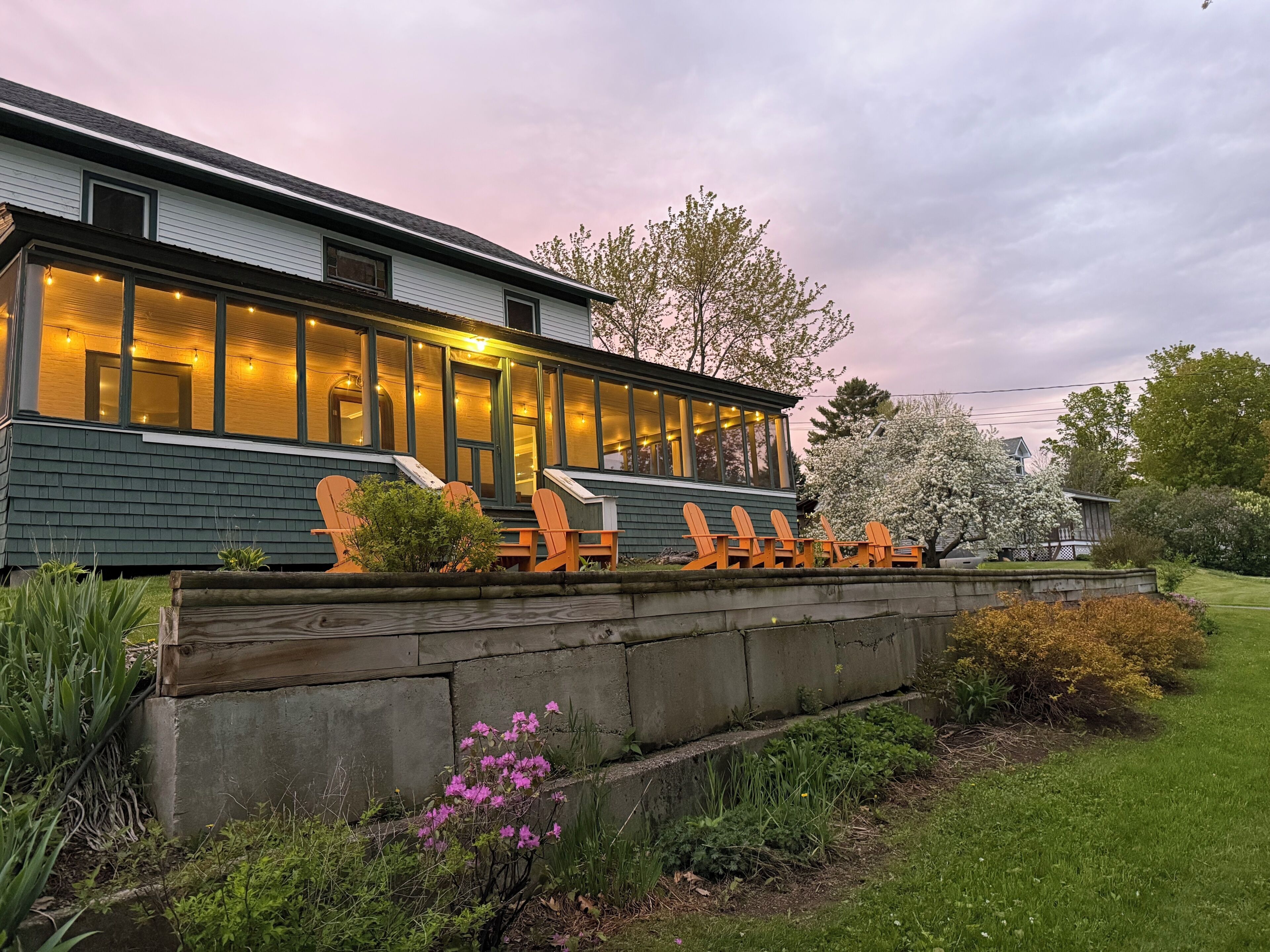 Un bâtiment moderne doté de grandes fenêtres, d’une terrasse avec des chaises en bois et d’un jardin bien entretenu.