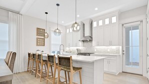 Una cocina moderna con armarios blancos, una isla central, taburetes de bar, iluminación colgante y vistas a través de un ventanal.