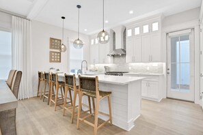 A modern kitchen with white cabinetry, a central island, bar stools, pendant lighting, and a view through a glass door. - Luxury villa w/ community pool & hot tub (Miramar Beach)