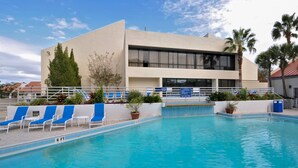 Ein Swimmingpool mit blauen Liegestühlen, ein Gebäude mit großen Fenstern und Palmen.