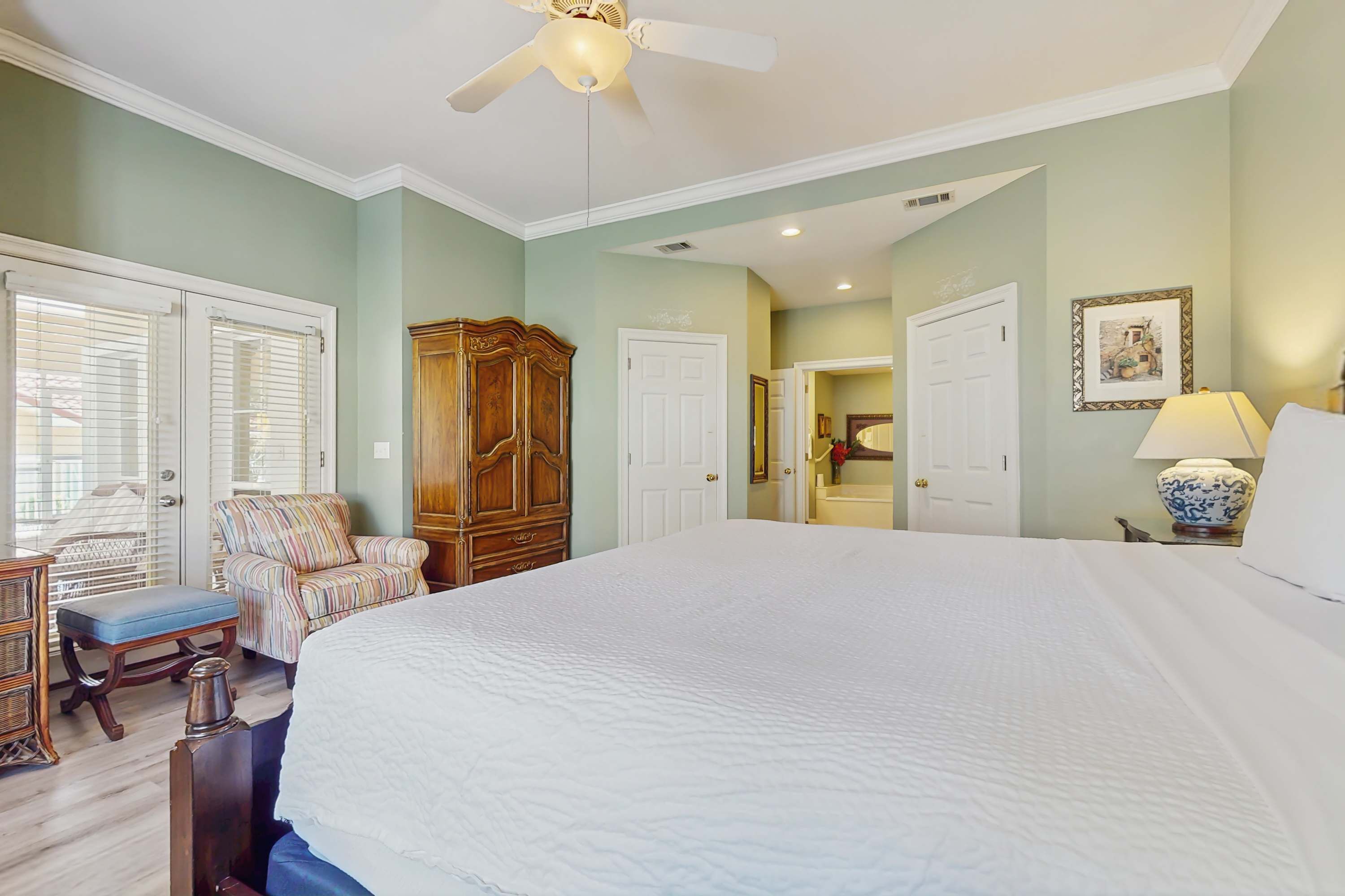 A bedroom with a large bed, a ceiling fan, a wooden wardrobe, a striped armchair, and a bedside table with a lamp.