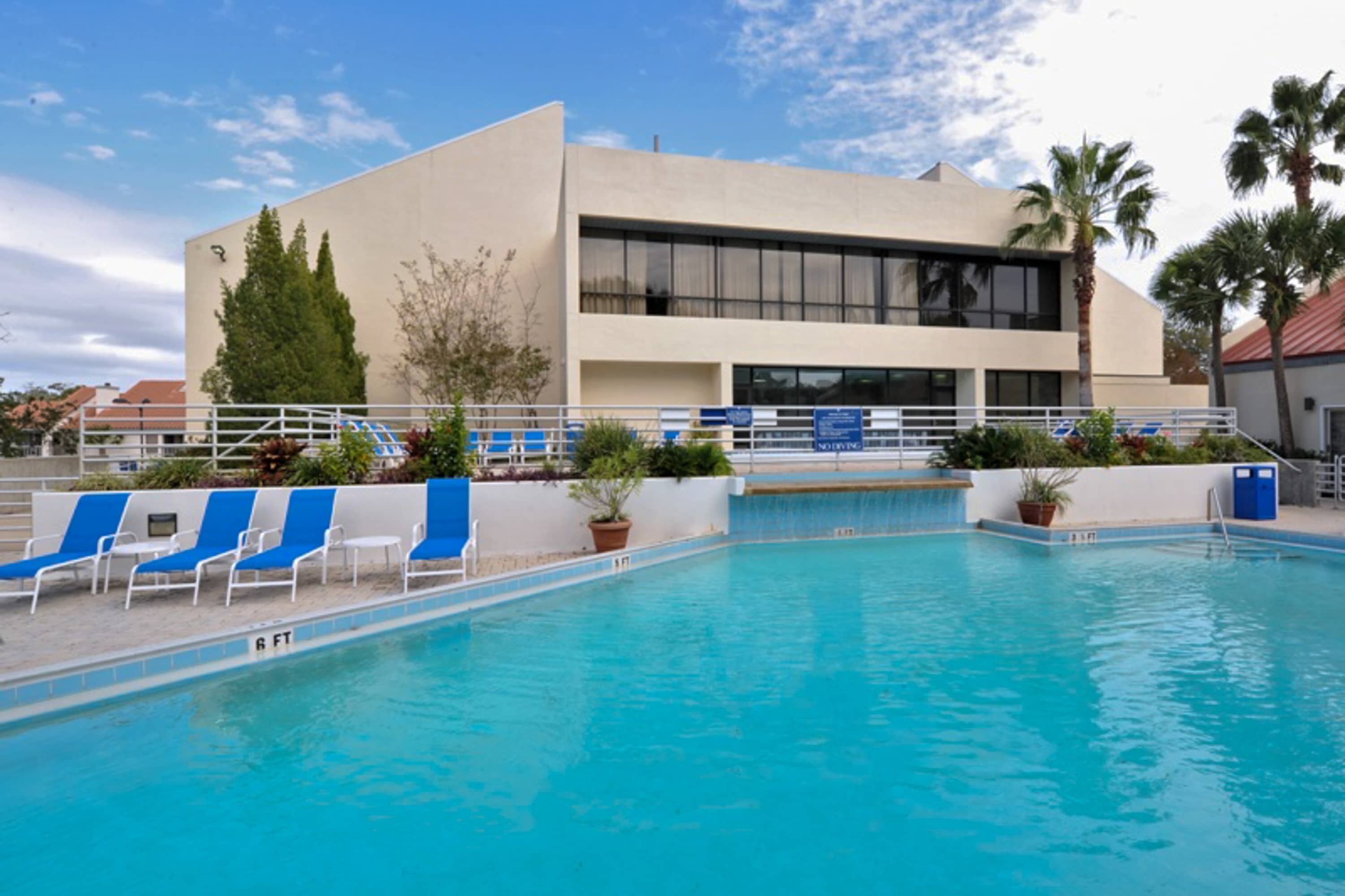 A swimming pool with blue chairs, a building with large windows, and palm trees.