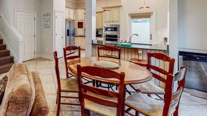 A dining area with a round table and wooden chairs, a kitchen with white cabinets and stainless steel appliances, and a staircase leading to another level. - 3BR villa, steps to beach + Captiva pool (Miramar Beach)