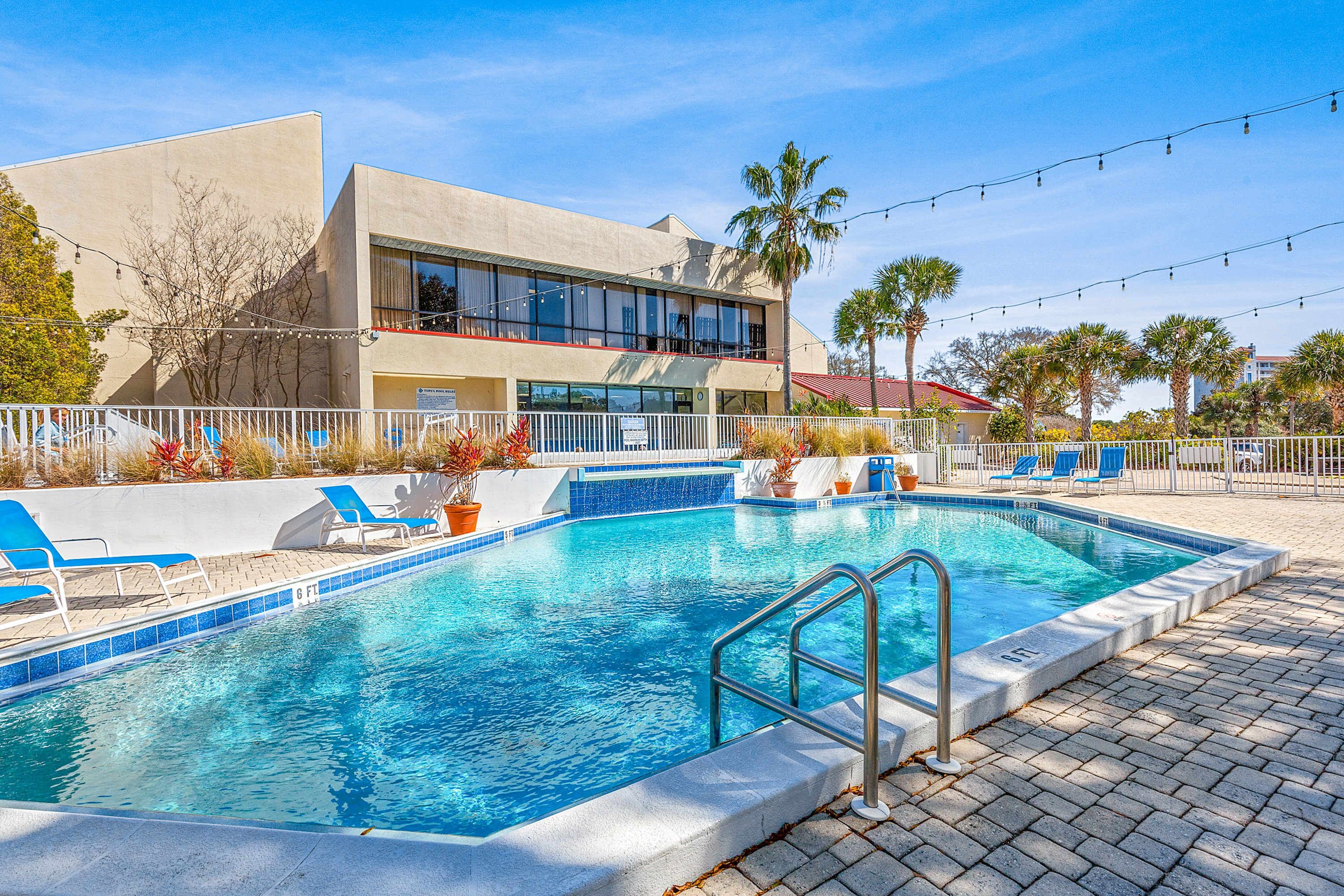 A swimming pool with lounge chairs, a fenced area, and a building with large windows and a red roof.