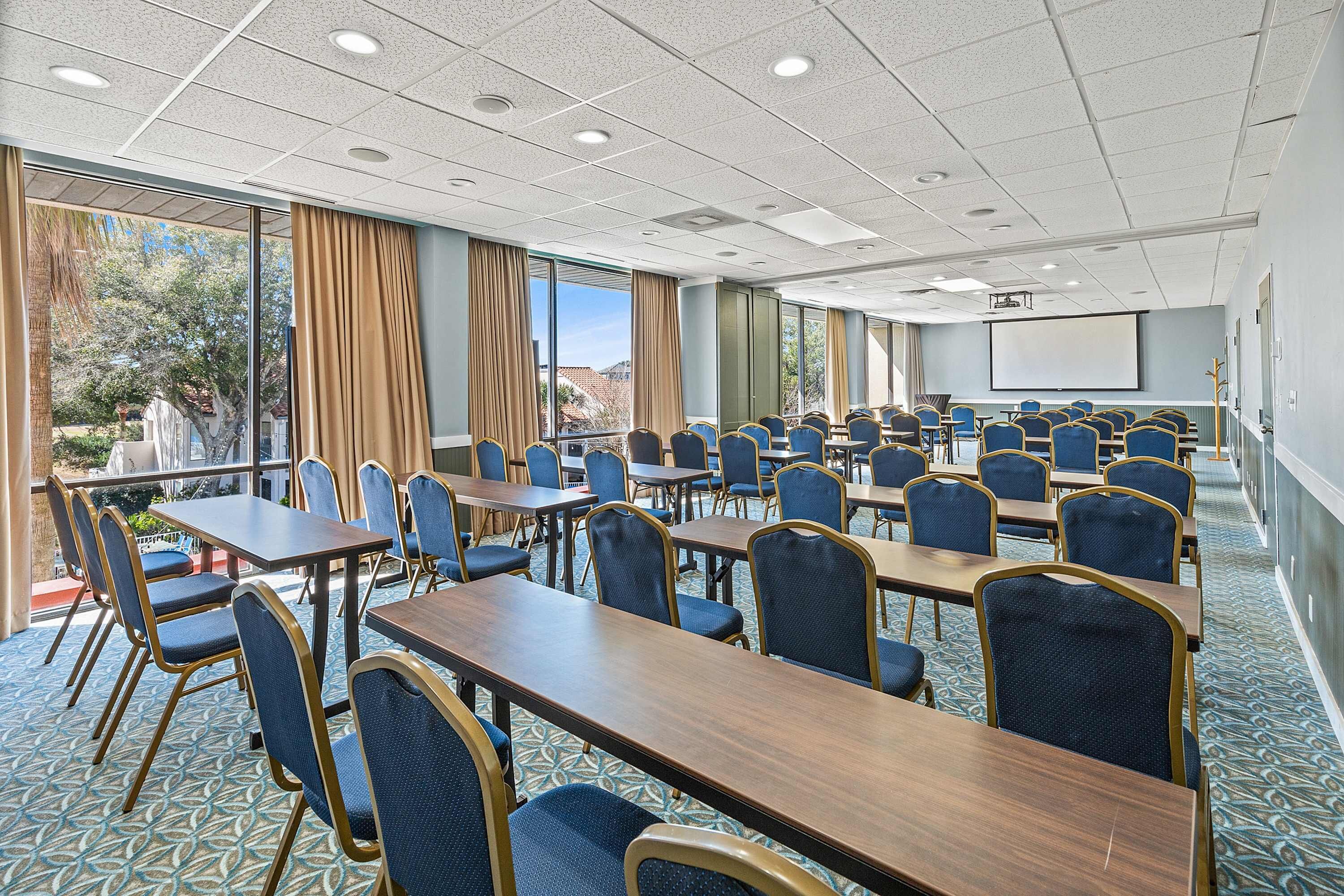 Une salle de conférence équipée de tables et de chaises, de grandes fenêtres et d’un écran de projection.