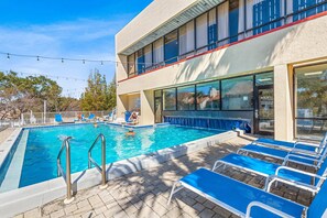 Una piscina con sillones reclinables azules, un área junto a la piscina y un edificio con ventanas amplias y un balcón.