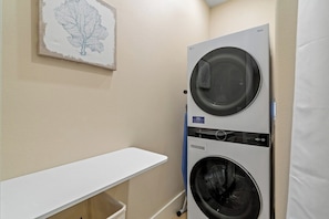 Un cuarto de lavandería con lavadora y secadora apiladas, un estante blanco y un cuadro con hojas en la pared.