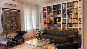 Ein Wohnzimmer mit einer Ledersofa, einem Glastisch, einem schwarzen Sessel, einem Bücherregal voller Bücher und einem gerahmten Bild an der Wand.