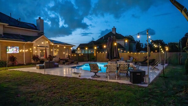 A backyard with a swimming pool, outdoor furniture, string lights, and a TV screen.