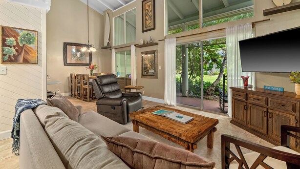 A spacious living room with a leather sofa, a wooden coffee table, and a TV on a wooden cabinet. There is a dining area with a table and chairs, and a large window offering a view of greenery outside. - (P9B) 2Br Princeville Retreat, Pool, AC, Wifi (Princeville)