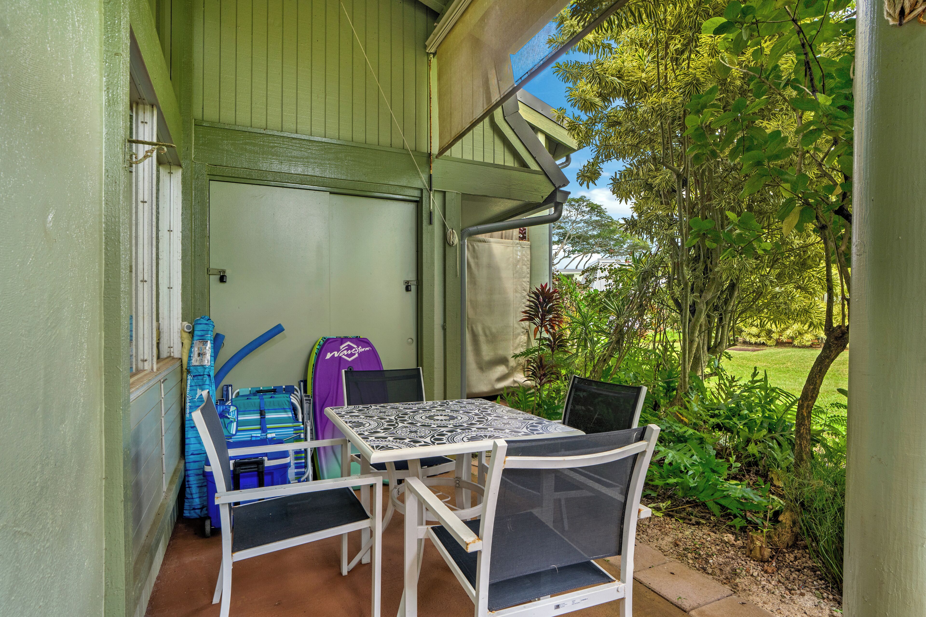 A covered patio with a table and chairs, surrounded by greenery and a view of the outdoors.