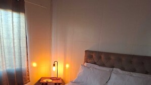 A neatly made bed with white linens and two folded towels, a bedside lamp, and a window with curtains. - Renovated house • Air conditi• Garage • BR-116 • Route to the beaches of SC (Guaíba)