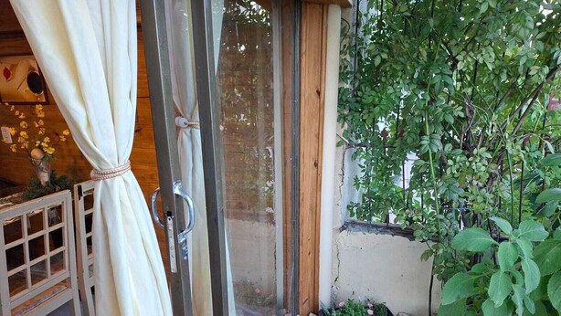 A wooden door with a glass panel, a small garden with plants and a pink pot, and a white curtain partially drawn. - Renovated house • Air conditi• Garage • BR-116 • Route to the beaches of SC (Guaíba)
