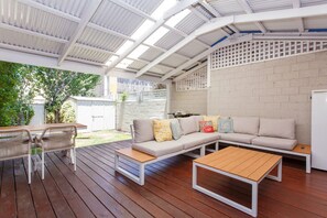 A covered patio with a wooden deck, a white sofa with cushions, a small table, and a white refrigerator. - Ocean River Rest - North Fremantle (North Fremantle)