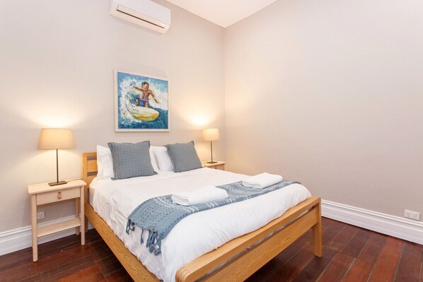 A neatly made bed with a blue and white blanket, two bedside lamps, and a painting of a child surfing on the wall. - Ocean River Rest - North Fremantle (North Fremantle)