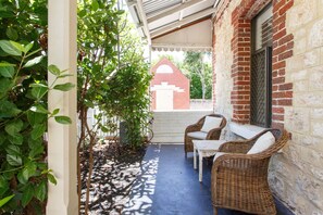 A covered outdoor seating area with wicker chairs and a table, surrounded by greenery and a brick wall. - Ocean River Rest - North Fremantle (North Fremantle)