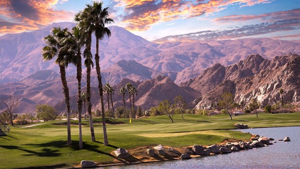Un campo de golf con palmeras, un lago y montañas al fondo.