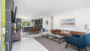 Una sala de estar moderna con un sofá, una mesa de centro y sillas, una zona de cocina con comedor, y un cuadro grande en la pared.
