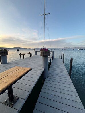 Eine Holzstege ragt in ruhiges Wasser, im Hintergrund Boote, eine hölzerne Bank und ein Blumenbeet.