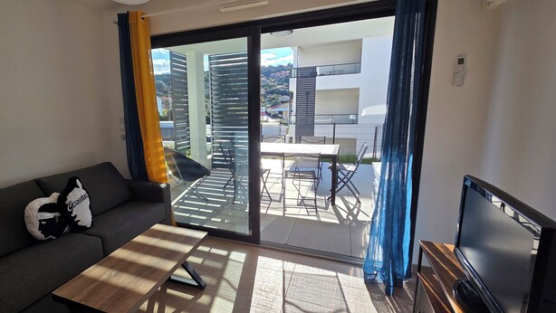 A modern living room with a sofa, a coffee table, a TV, and a sliding glass door leading to an outdoor area with a table and chairs. - Nice and cozy T2, near beaches, Terra Doru B02 (Propriano)