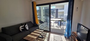 Un saló modern amb un sofà, una taula de centre, una televisió i una porta corredissa de vidre que dona a una zona exterior amb una taula i caires.