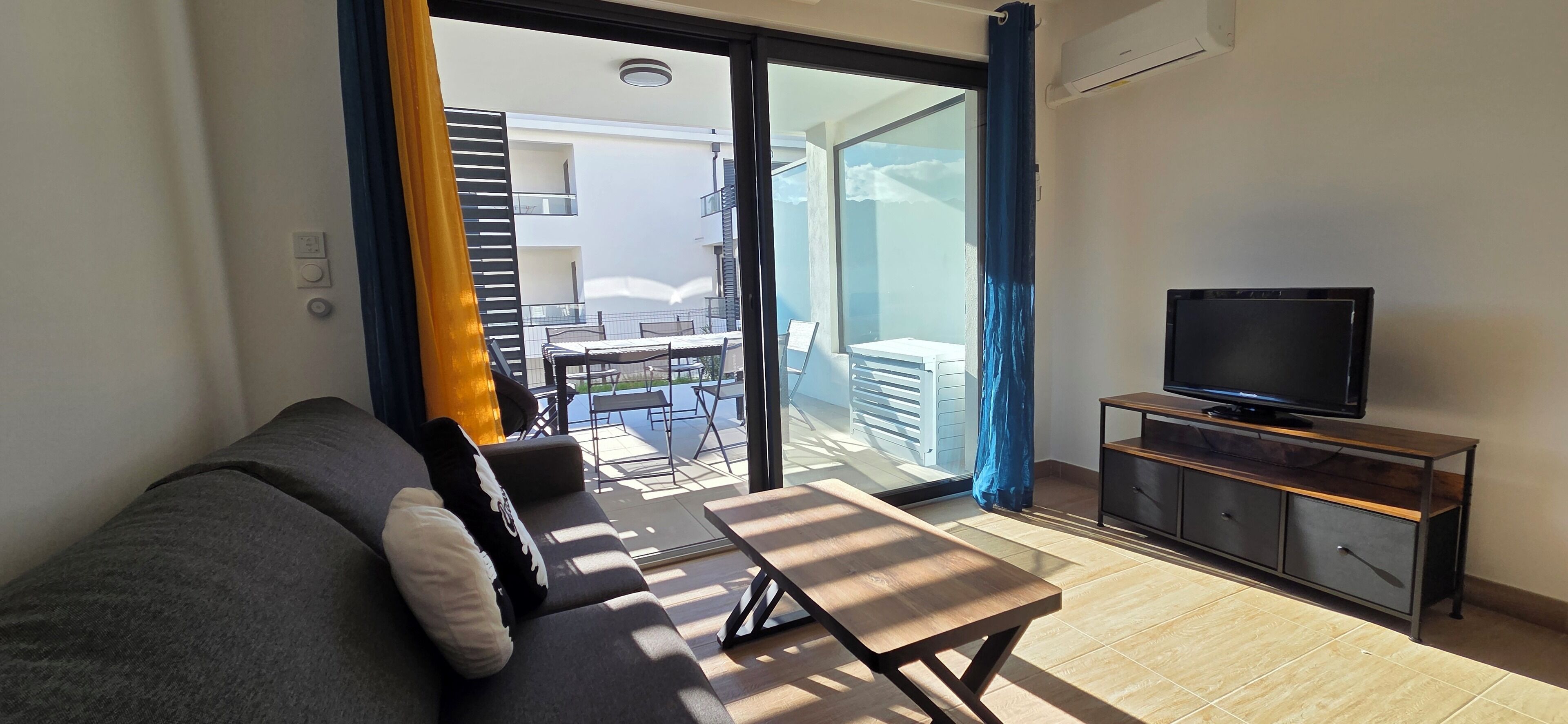 Uma sala de estar moderna com sofá, televisão sobre suporte de madeira, e varanda com mesa e cadeiras.