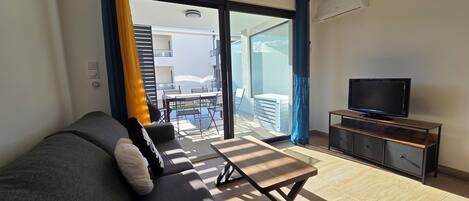 Uma sala de estar moderna com sofá, televisão sobre suporte de madeira, e varanda com mesa e cadeiras.