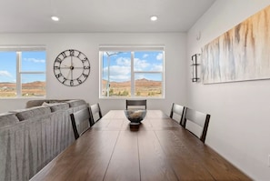 Une salle à manger avec une table en bois, des chaises, une horloge et une vue sur un paysage désertique, visible à travers de grandes fenêtres.