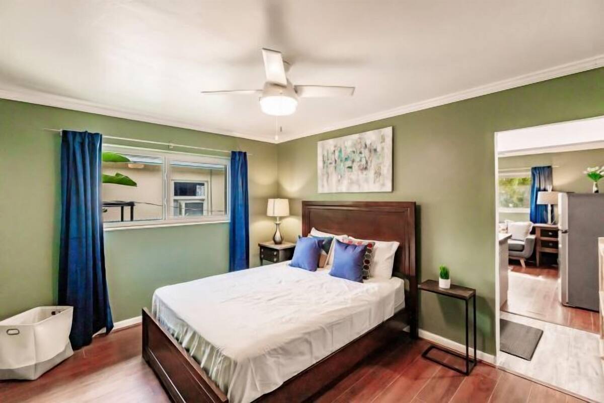 Un dormitorio con cama, ventilador de techo, mesita de noche, ventana con cortinas y vista al área de estar contigua.
