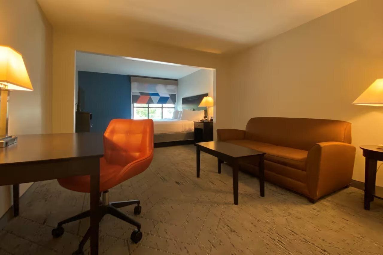 A hotel room with a bed, a desk, an orange chair, a brown sofa, and two lamps.