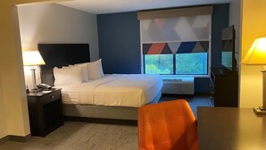 A hotel room with a bed, a desk, a chair, a TV, and a window with a view of greenery. - Holiday Inn Express & Suites RALEIGH WEST - LENOVO CENTER by IHG (Meredith)