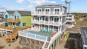 A multi-story beach house with a pool, deck chairs, and a water tower in the background. - Clear Skies: Spacious 7-Bedroom Oceanfront Retreat in Oak Island, NC, perfect for up to 20 guests. (Oak Island)