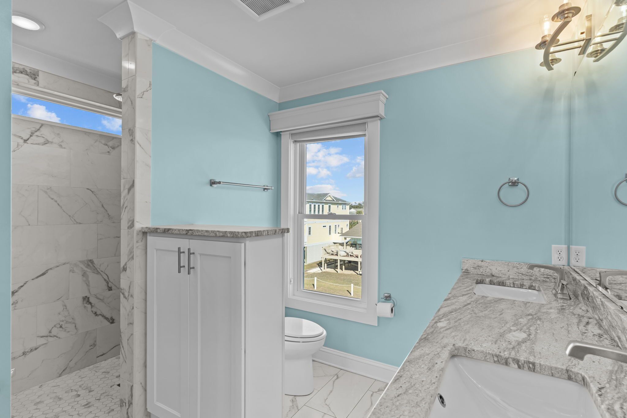 A bathroom with a marble shower, a toilet, a sink with a vanity, and a window with a view of a house.