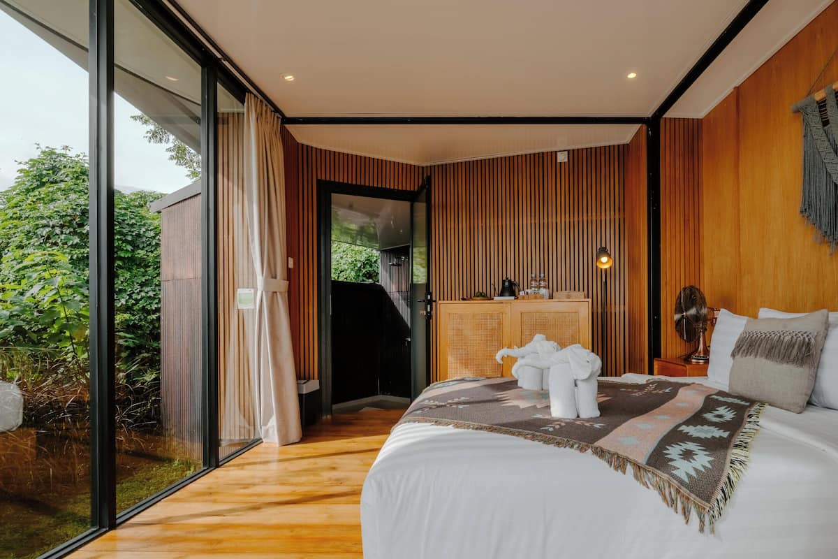 Un dormitorio moderno con una cama grande, paneles de madera, vistas a través de un ventanal y un mueble bajo con objetos encima.