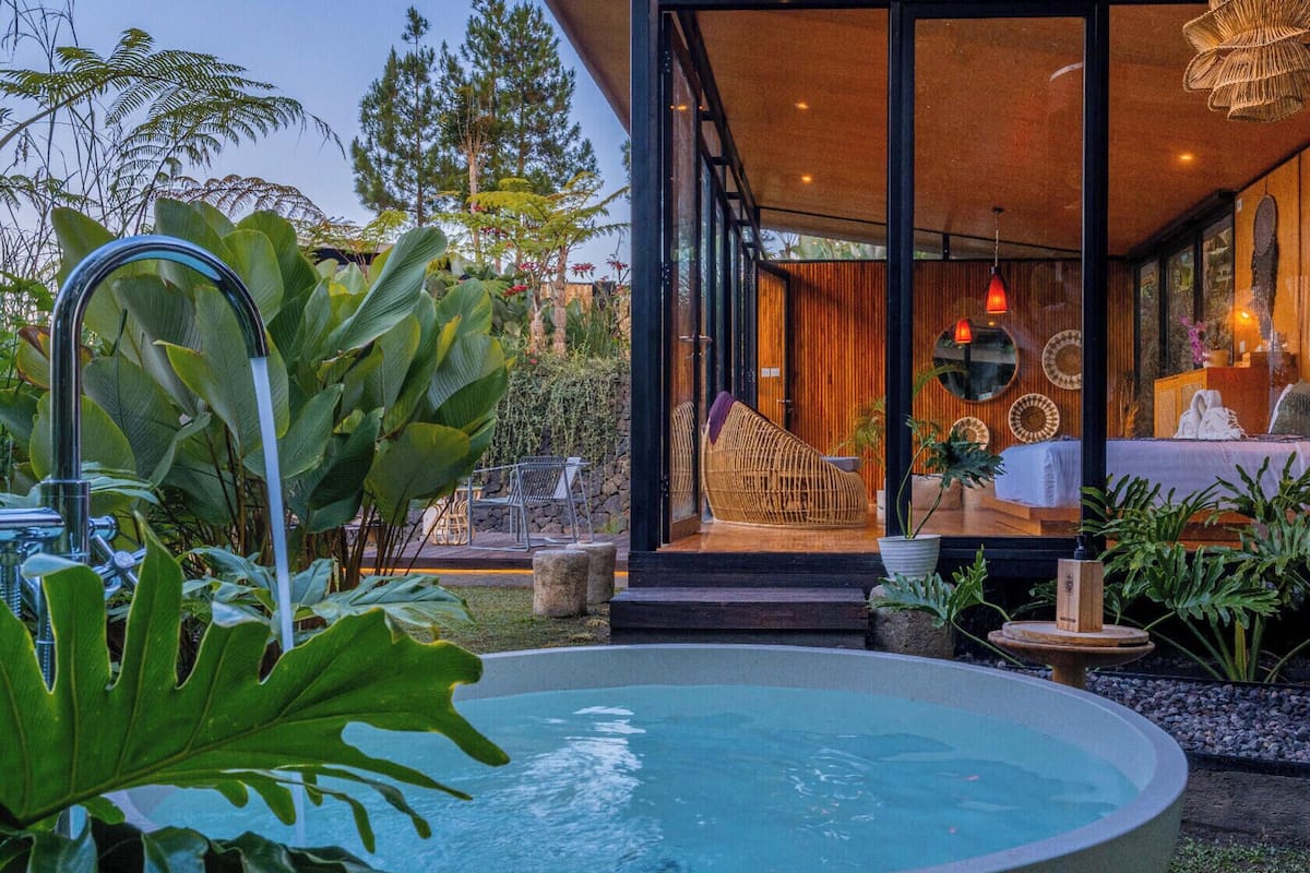 Una casa moderna con piscina, una terraza de madera y un dormitorio con paredes de cristal que incluye una cama y un sofá.