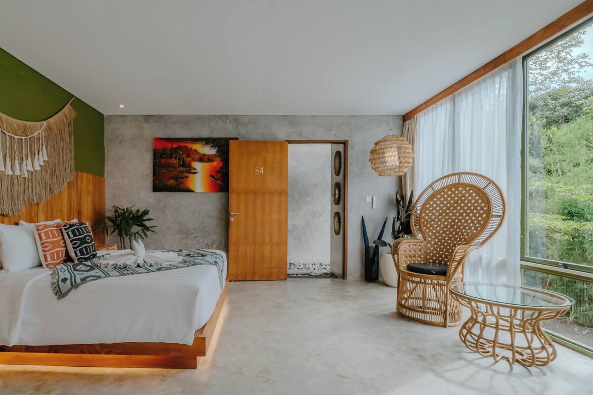 Un dormitorio moderno con cama, un ventanal grande, una silla de ratán y un cuadro en la pared.