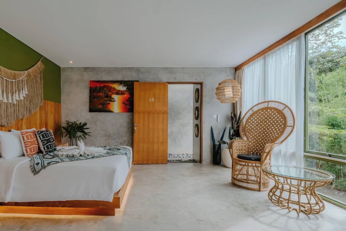 Un dormitorio moderno con cama, un ventanal grande, una silla de ratán y un cuadro en la pared.