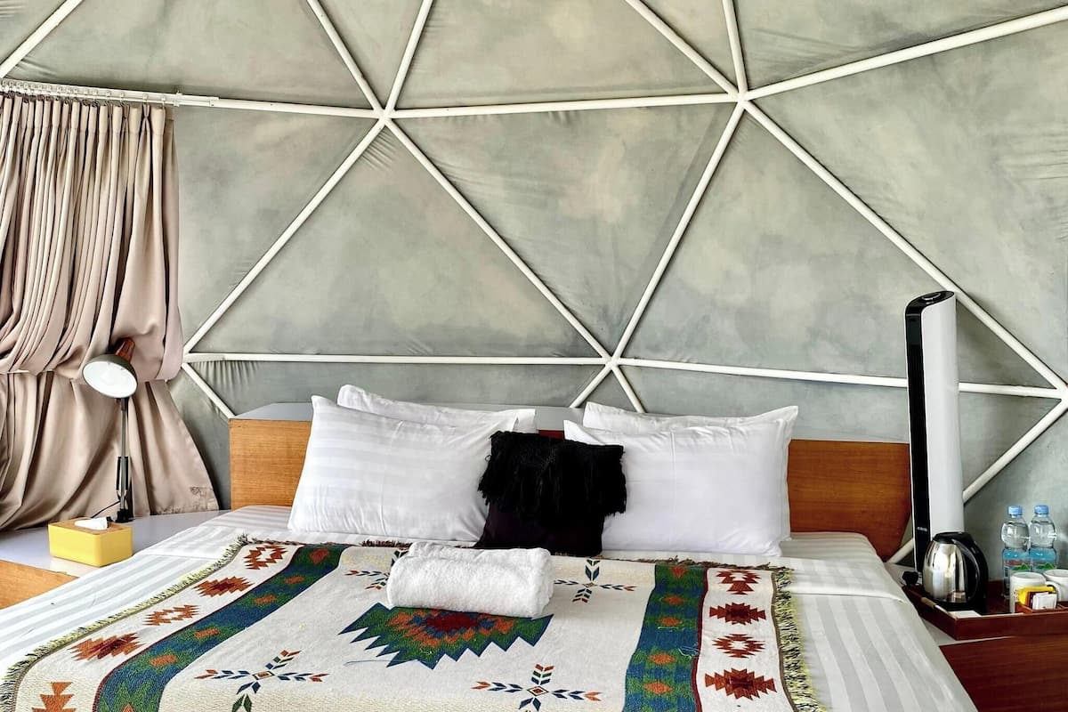 Una habitación con techo de diseño geométrico, una cama con ropa blanca, una lámpara de noche y una botella de agua sobre la mesita.