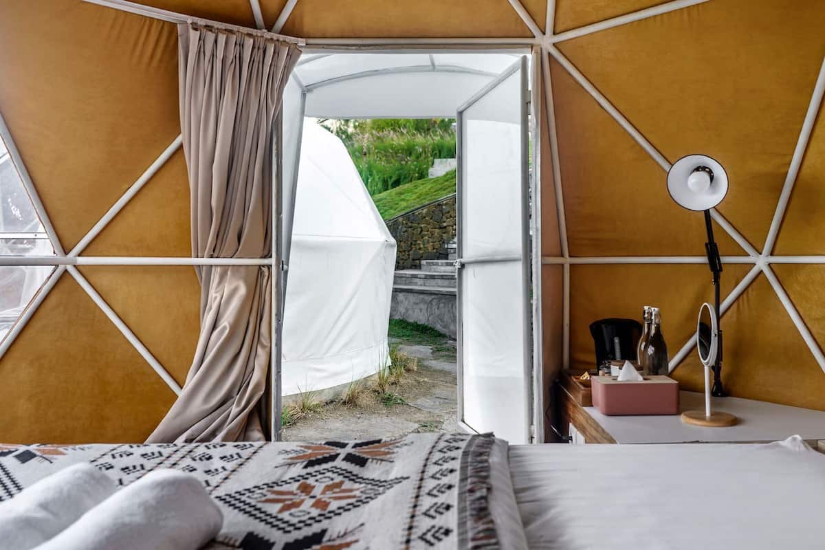 Alojamiento en tienda de campaña con cama, mesita de noche con lámpara y vistas a la vegetación.