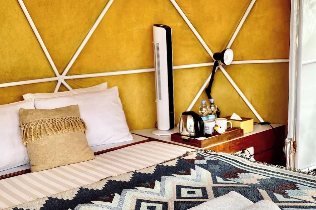 Una cama con almohadas blancas y beige, una manta estampada, una mesita de noche con una tetera, una botella de agua y una taza.