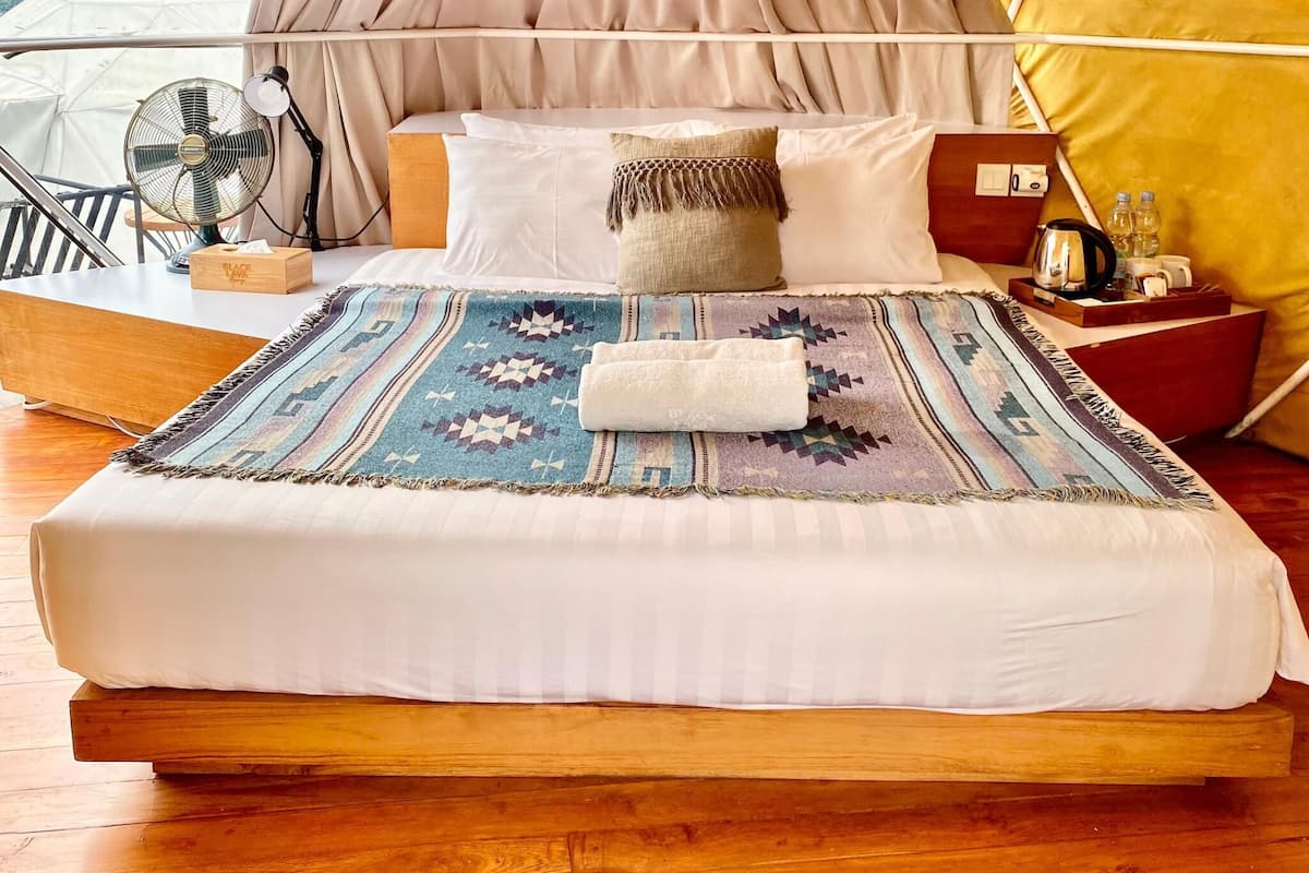 Una cama con una manta estampada, almohadas, una mesita de noche de madera con una tetera y un ventilador.