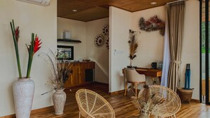 A room with a wooden floor, a dining area with wicker chairs, a kitchen in the background, and a large window with curtains. - Black Lava Camp Kintamani (Kintamani)