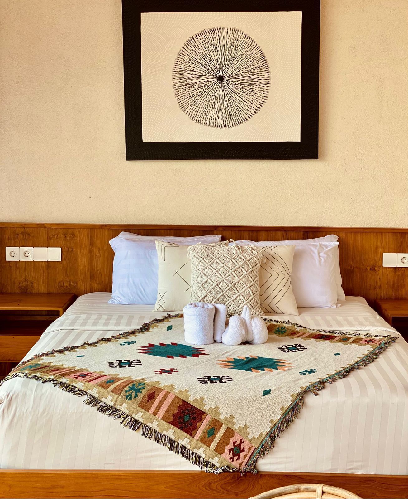 Una cama con una manta estampada, almohadas blancas y una obra enmarcada en la pared.