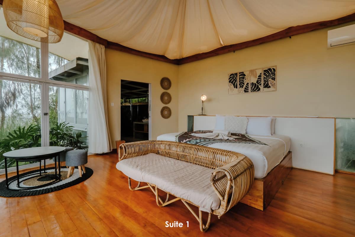 Un dormitorio con una cama grande, un banco de mimbre, una mesa redonda y vistas a zonas verdes al exterior.
