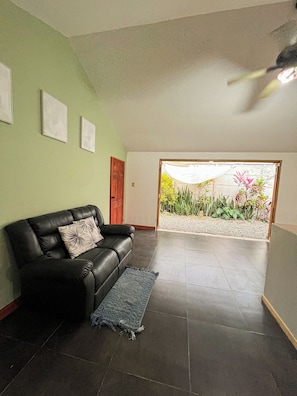 A spacious living room with a black leather sofa, a blue rug, and a door leading to a garden with plants and a gravel path. - Casa Miel: Sweet Getaway in Paradise (Puerto Jiménez)