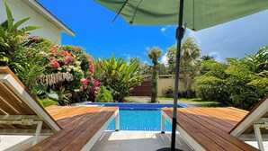 Una piscina con sillones de madera y una sombrilla, rodeada de exuberante vegetación y un cielo azul despejado.